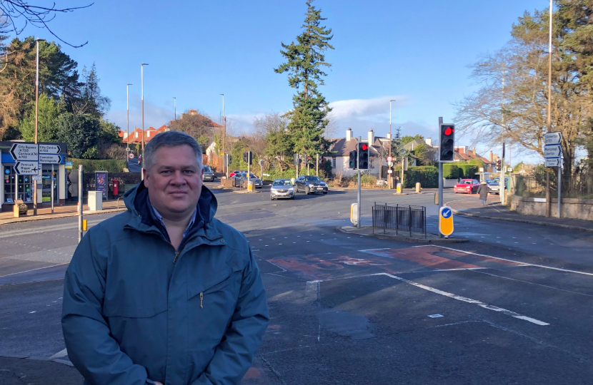 Neil is standing in front of a junction, smiling at the camera. 