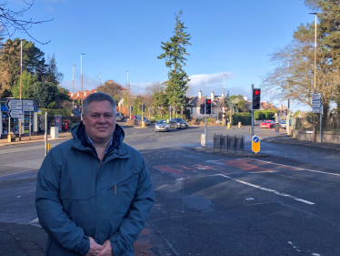 Neil is standing in front of a junction, smiling at the camera. 